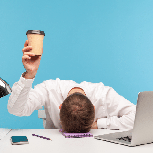 Man hiolding up a coffee cup while his head is on his desk, exhausted and burned out
